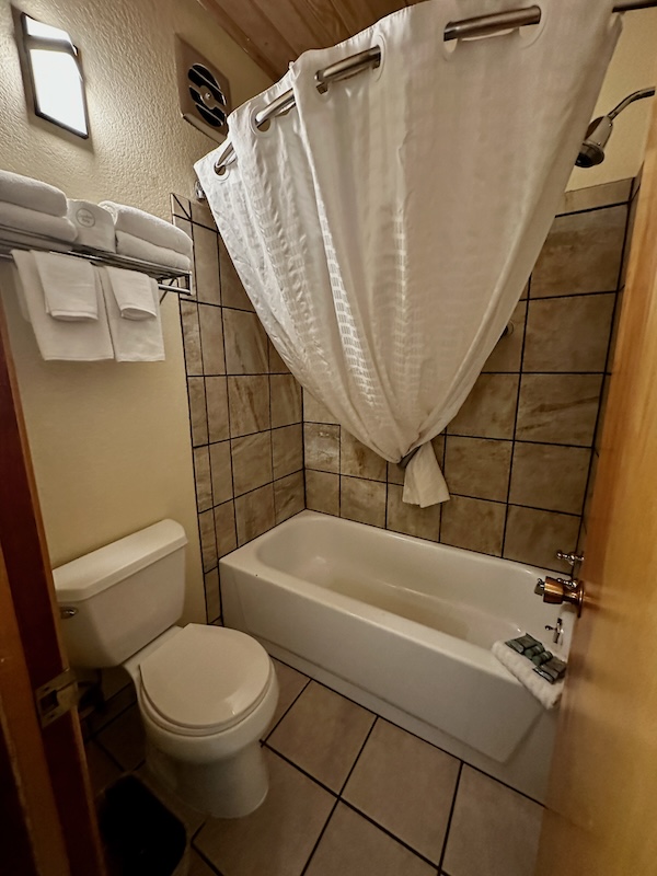 A bathroom with tiled floors and walls featuring a bathtub with a white shower curtain, a toilet, and a towel rack holding multiple folded towels.