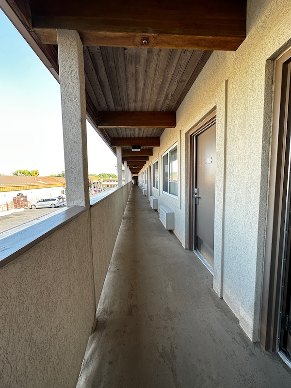 A long, open-air corridor on the second floor of the motel with doors leading to guest rooms. The corridor features wooden beams and a view of the parking lot below.