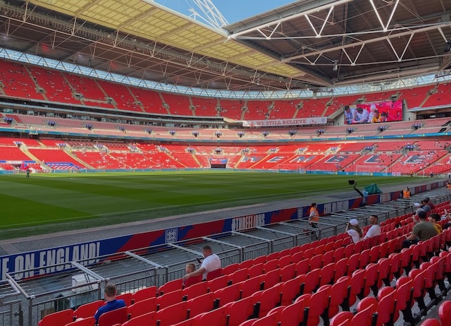 Sunny day at Wembley Stadium with early arriving fans taking their red seats. The large screen displays a football match highlight, under the iconic Wembley arch and a clear blue sky, emphasizing the anticipation of an upcoming match.