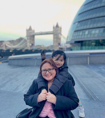 A woman with glasses, wearing a black coat, carrying a child piggyback style, with the iconic Tower Bridge in the background on a cloudy day.