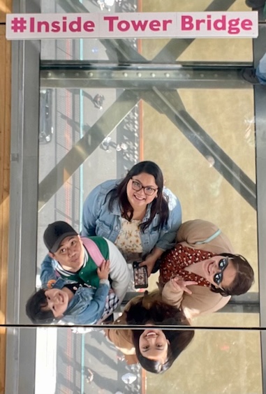 A group of people, including four adults and a child, joyfully smiling above a transparent glass floor with the hashtag #Inside Tower Bridge visible, enjoying one of the best London attractions for families at the Tower Bridge Exhibition in London.