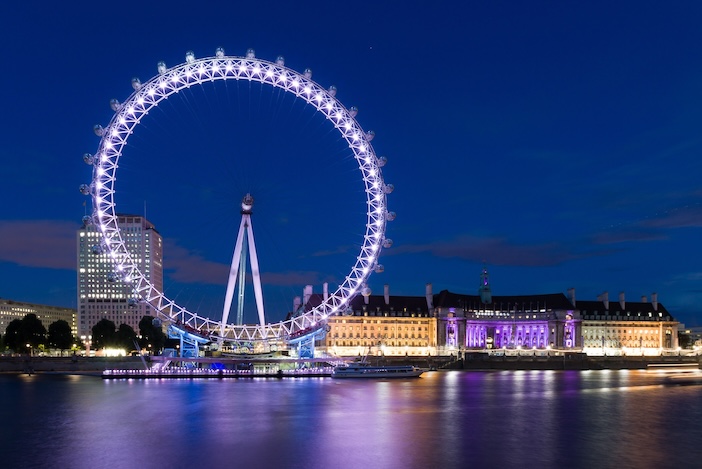 The London Eye illuminated against the evening sky, with the River Thames in the foreground reflecting city lights. The nearby County Hall is also lit up, adding to the vibrant nighttime allure of this popular London attraction.
