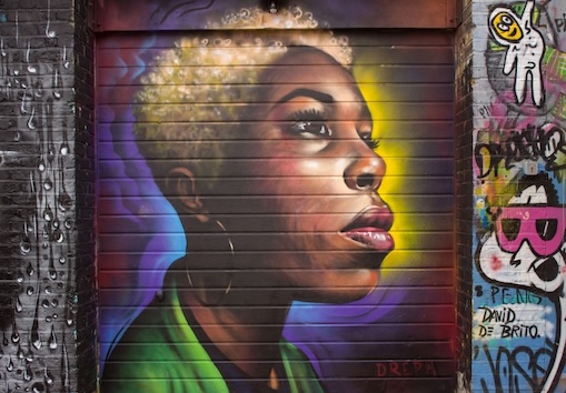A vivid mural on Shoreditch's street depicts the profile of a woman with striking features and a spectrum of colours, alongside urban tags and expressive graffiti.