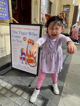 A joyful child in a purple dress and tiger-striped headband roars playfully beside a promotional poster for 'The Tiger Who Came to Tea' theatre show, describing it as the 'PURRFECT SUMMER TREAT!