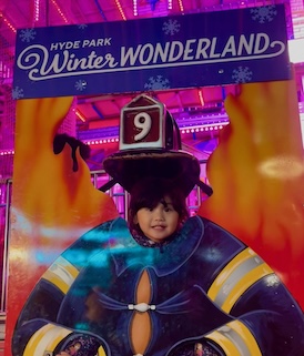 A child smiles while peeking through a cutout photo board at Hyde Park's Winter Wonderland, with the festive event's signage above.