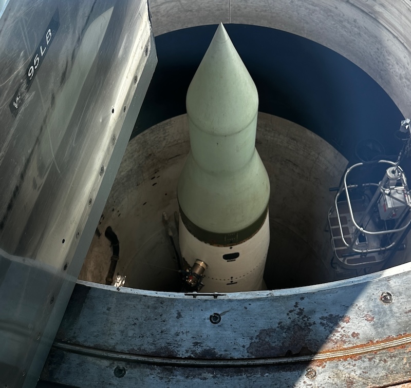 A view looking down into a missile silo at the Minuteman Missile National Historic Site, showing a Minuteman missile in its launch position with the tip pointed upward. The surrounding structure is made of concrete, and a metal ladder is visible on the right side.