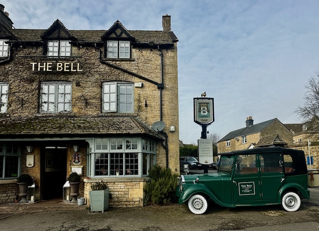 The Bell, a rustic stone pub-style hotel in Stow-on-the-Wold, features a classic green vintage car parked outside under a hanging sign, showcasing its traditional countryside charm.