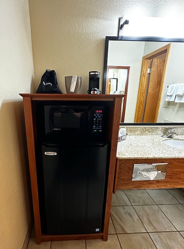 A mini fridge and microwave unit topped with a coffee maker, ice bucket, and a black bag labeled "Dryer" next to a bathroom vanity with a granite countertop and sink.