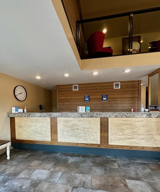 Best Western Plains Motel, Wall, South Dakota: The lobby area with a wooden reception desk, a granite countertop, and a clock on the wall showing the time. Above the desk is an open second floor with red chairs.