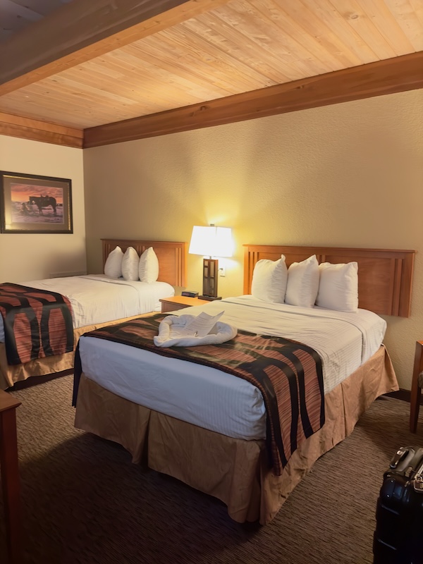 A hotel room with two double beds, wooden headboards, and a wooden ceiling. Each bed is neatly made with white linens and several pillows. A painting of horses is visible on the wall. A black luggage is seen in the corner.