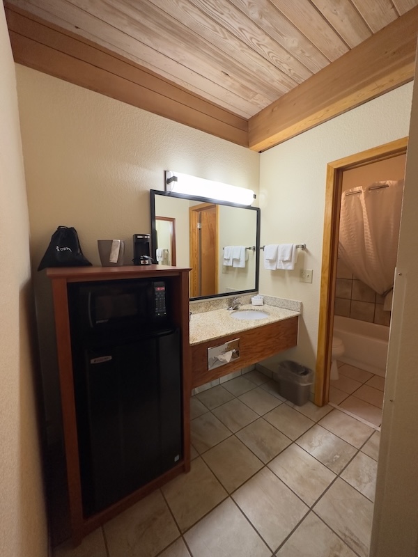 Best Western Plains Motel Review - A bathroom vanity area featuring a granite countertop, a sink, a large mirror, and a small refrigerator with a microwave on top. An ice bucket, coffee maker, and a black bag labeled "Dryer" are on the countertop.