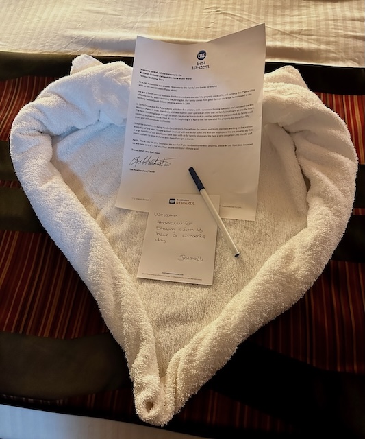 A welcome note on a bed at Best Western Plains Motel, shaped into a heart with towels. The note, signed by the owner, expresses appreciation for the guest's stay and includes a handwritten message: "Welcome, thanks for staying with us, have a wonderful day. Sincerely, Jolene.