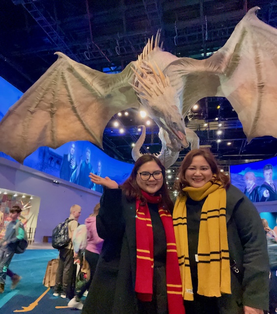 Two women smile in front of a life-sized dragon model from the Harry Potter series, each wearing long coats with Gryffindor and Hufflepuff scarves as Harry Potter costumes. The dramatic scene captures the magical atmosphere with the dragon’s wings spread wide above them.
