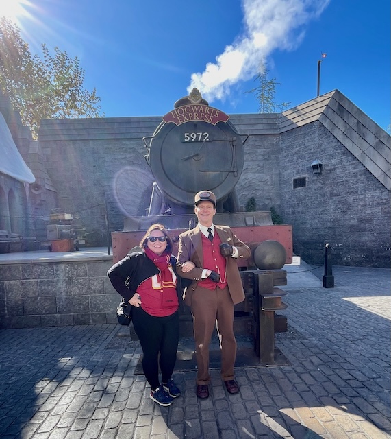 A woman dressed in a red and gold Gryffindor Harry Potter costume poses cheerfully beside the Hogwarts Express conductor in front of the train marked with the number 5972. The iconic steam engine is parked in a cobblestone courtyard under a clear blue sky.
