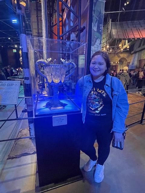 A smiling woman stands next to the Triwizard Cup displayed in a glass case at a Harry Potter exhibit. She wears a denim jacket over a Hogwarts-themed shirt, highlighting her love for the series while posing with the iconic magical trophy.