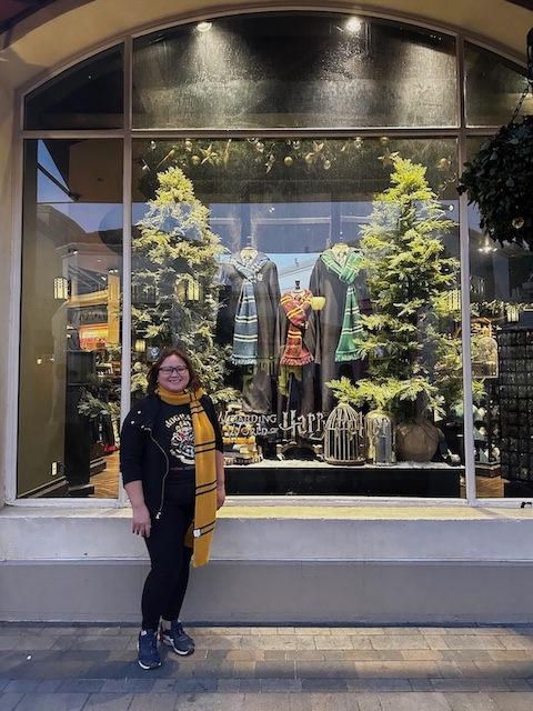 A smiling woman stands in front of a store window display at The Wizarding World of Harry Potter, wearing a black outfit with a yellow Hufflepuff scarf and Hogwarts-themed shirt. Behind her, mannequins are dressed in Hogwarts robes from Ravenclaw, Gryffindor, and Slytherin, showcasing detailed Harry Potter costumes framed by faux pine trees.