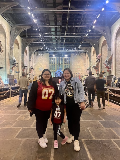 Three people, including a young child, pose inside a detailed replica of the Hogwarts Great Hall, wearing matching Gryffindor-themed Harry Potter costumes. The stone hall features floating candles, gothic arches, and banners hanging in the background, recreating a magical scene from the films.
