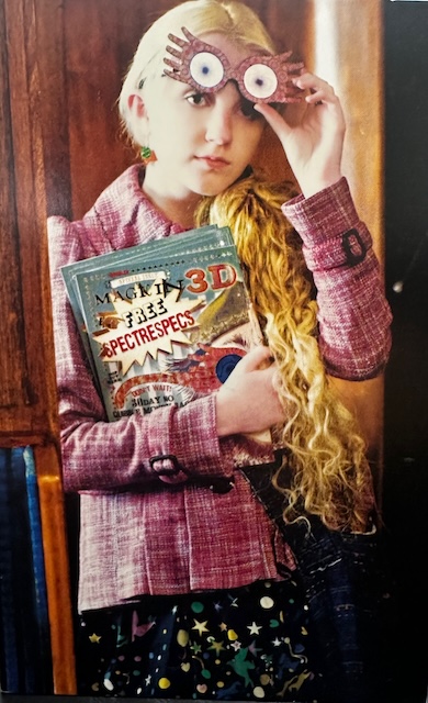 Luna Lovegood poses with pink Spectrespecs glasses and a copy of The Quibbler. She wears a pink jacket and patterned skirt, embodying Luna’s quirky style.