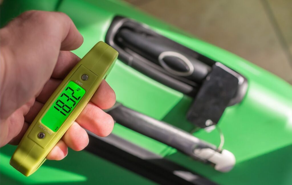 Hand holding a digital luggage scale reading 18.32 kg, with a green suitcase handle visible in the background.