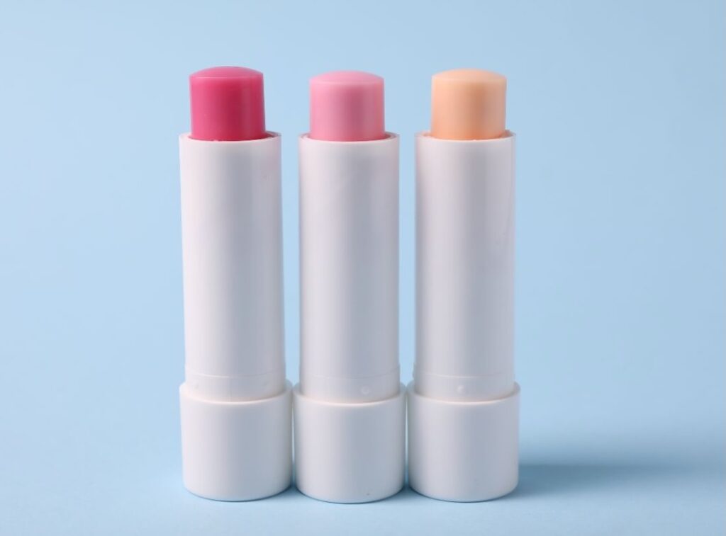 Three white lip balm tubes with caps removed, showing red, pink, and peach-colored balms against a blue background.