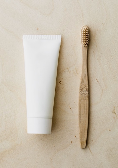A bamboo toothbrush and a minimalist white toothpaste tube on a light wooden surface. Sustainable oral care items that make thoughtful travel stocking stuffers.
