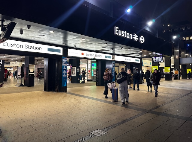 Entrance to London Euston Station at night with travellers walking past illuminated “Euston Station” signs and rolling luggage through the busy terminal. One of the main departure points travellers use when researching how to get to Harry Potter Studios from central London.