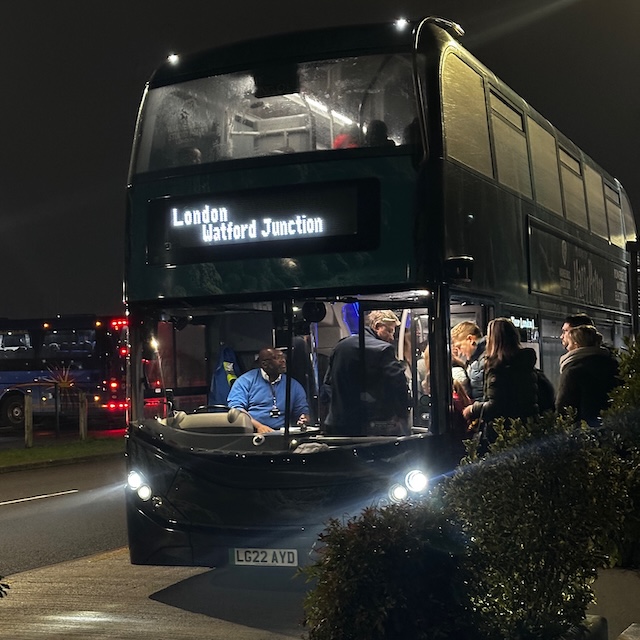 Harry Potter Studio Tour shuttle bus at night with passengers boarding outside Warner Bros Studio Tour - The Making of Harry Potter waiting area to get to Watford Junction station under glowing headlights. The shuttle connection is one of the easiest transport options for first time visitors travelling to Harry Potter Studios London.