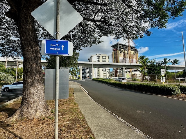 Blue sign reading "Bus To Waikiki" stands beside a sidewalk and road lined with trees, with Honolulu buildings visible in the distance. The photo shows a transit stop area that helps explain how to get to Pearl Harbor without a car.