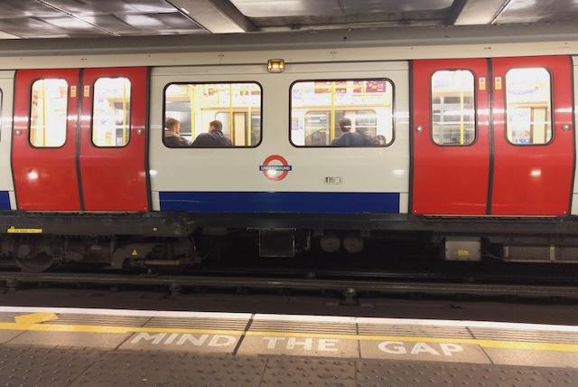 London Underground train stopped at a platform with passengers visible through the windows and the words "MIND THE GAP" painted along the platform edge. The photo illustrates part of the public transport journey for visitors researching how to get to Harry Potter Studios from Heathrow.