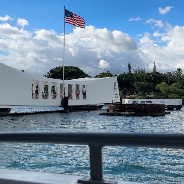 The white USS Arizona Memorial and the "USS Arizona BB 39" marker are viewed from a boat approaching the memorial beneath a waving American flag.